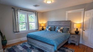 A bedroom with a bed, two bedside tables, a nightstand, a potted plant, and a window with curtains. - Stay Roanoke | Downtown Home + Greenway Access (Roanoke)
