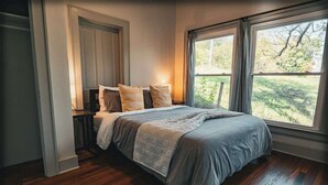 A bedroom with a large bed, wooden flooring, a window with a view of greenery, and a door leading to another room. - Stay Roanoke | Rustic Cabin Near McAfee Knob (Catawba)