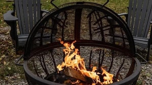 A fire pit with burning logs, surrounded by two Adirondack chairs, set on a gravel surface with a house and trees in the background. - Stay Roanoke | Rustic Cabin Near McAfee Knob (Catawba)