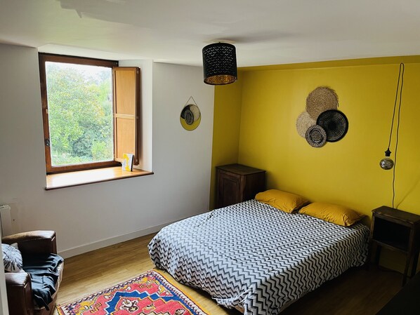 A bedroom with a large bed, a window with a view of greenery, a small table, and a chair.