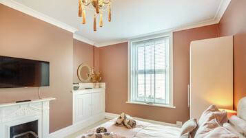 A bedroom with a fireplace, a television, a bed with pillows, a nightstand, and a window with blinds.