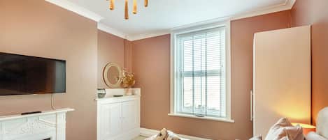 A bedroom with a fireplace, a television, a bed with pillows, a nightstand, and a window with blinds.