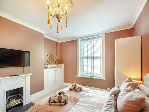 A bedroom with a fireplace, a television, a bed with pillows, a nightstand, and a window with blinds.