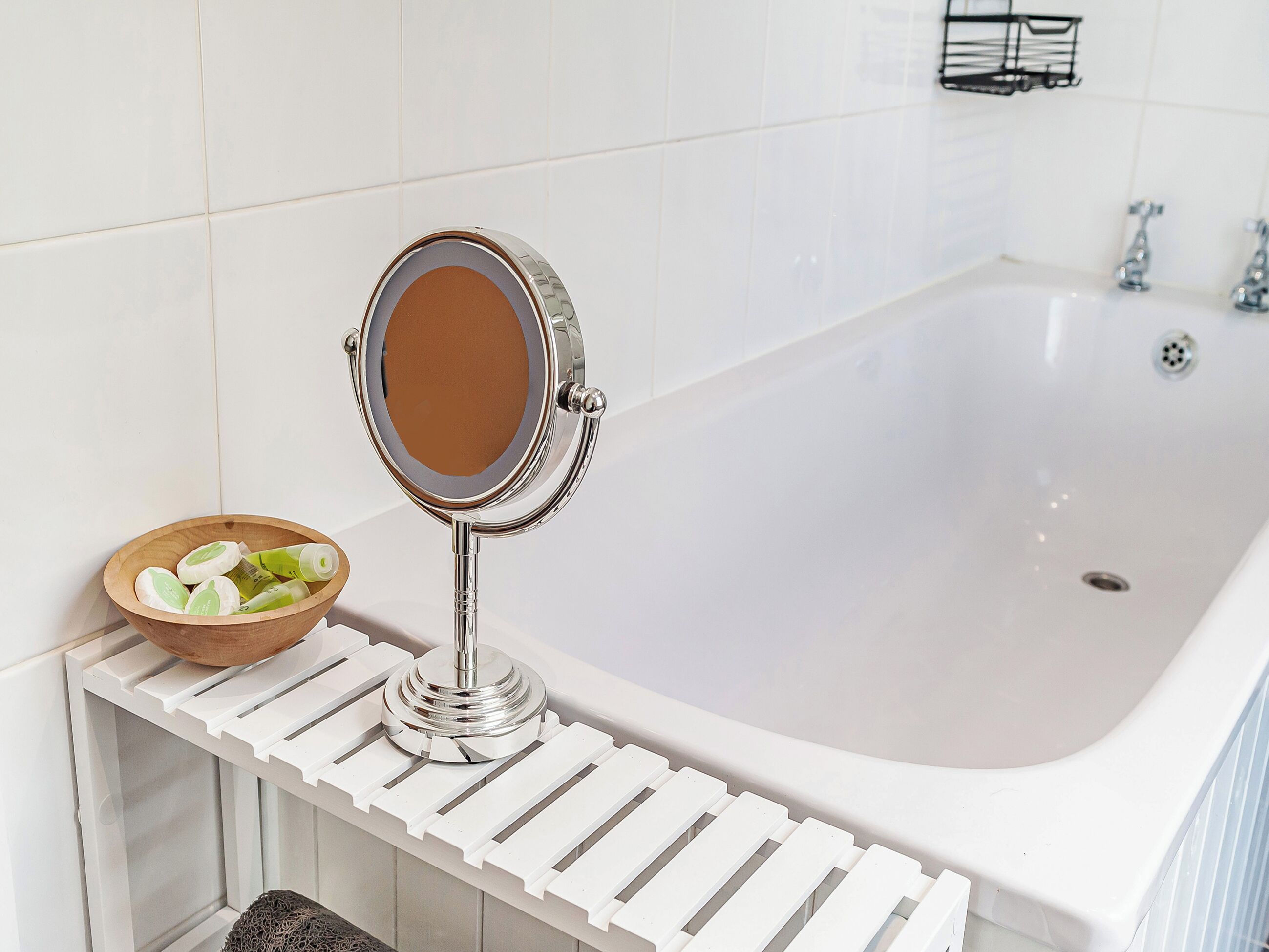 Une salle de bain avec une baignoire blanche, un miroir rond posé sur un support blanc, et un bol en bois contenant des objets verts, le tout sur une étagère blanche.