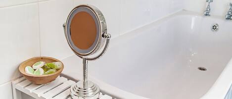 Une salle de bain avec une baignoire blanche, un miroir rond posé sur un support blanc, et un bol en bois contenant des objets verts, le tout sur une étagère blanche.