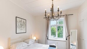 Una camera da letto con un letto, comodini, un lampadario, una finestra con tende, uno specchio e un quadro appeso al muro.
