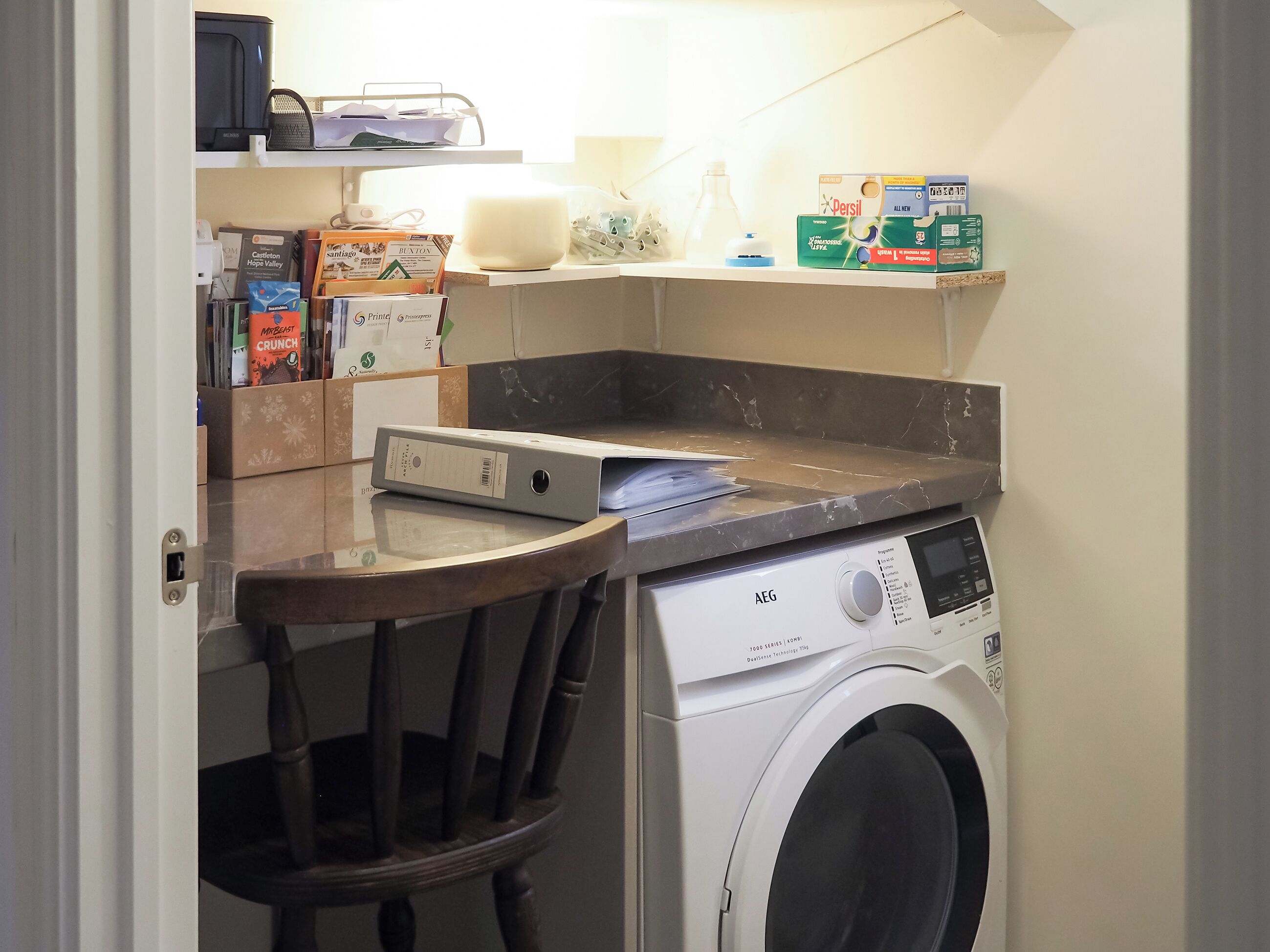 A compact laundry area with a washing machine, a small table, and shelves holding various items including boxes and a bottle.