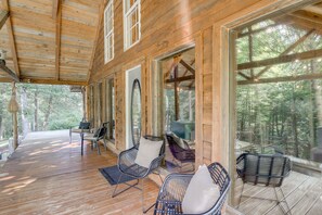 Una cabaña de madera con un porche cubierto, amueblada con sillas de metal y una mesa, con vistas a un bosque.