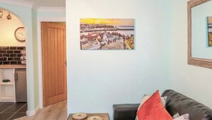 Een woonkamer met een zwarte leren bank, een houten salontafel met twee ronde houten objecten, een schilderij van een kustplaats, en een keuken in de achtergrond.