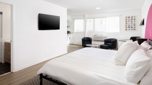 A modern hotel room with a large bed, a flat-screen TV mounted on the wall, a desk with a chair, and a view of the city through large windows. - Fenix Hotel Hollywood (Los Angeles)