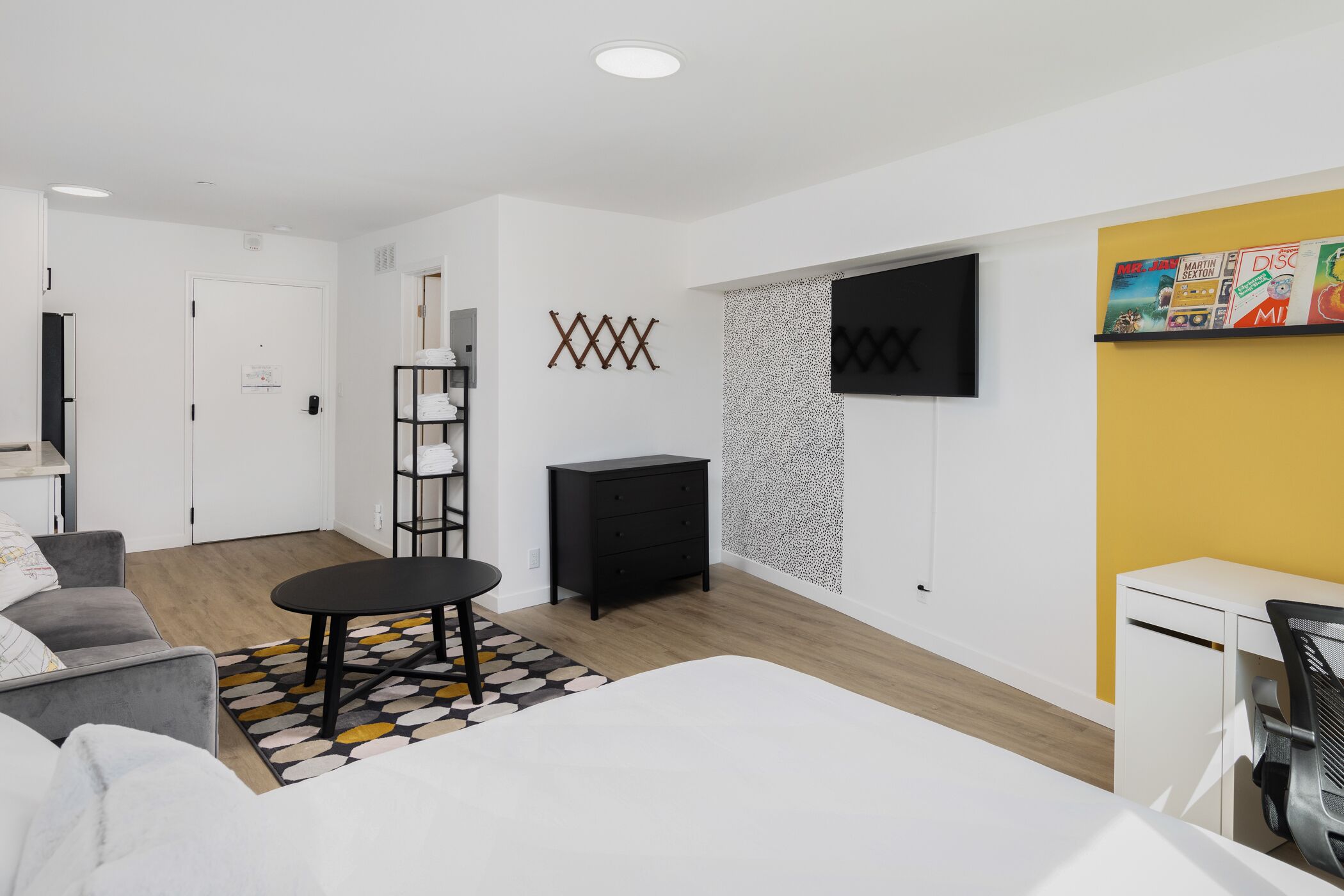 Une chambre moderne et bien éclairée, équipée d’un lit, d’un bureau, d’une chaise, d’une télévision et d’une étagère où sont disposés des objets décoratifs.