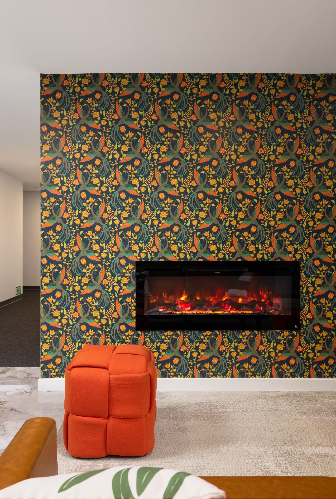 Ein Zimmer mit gemustertem Wandteppich, einem Kamin und einem modernen, orangefarbenen Pouf.