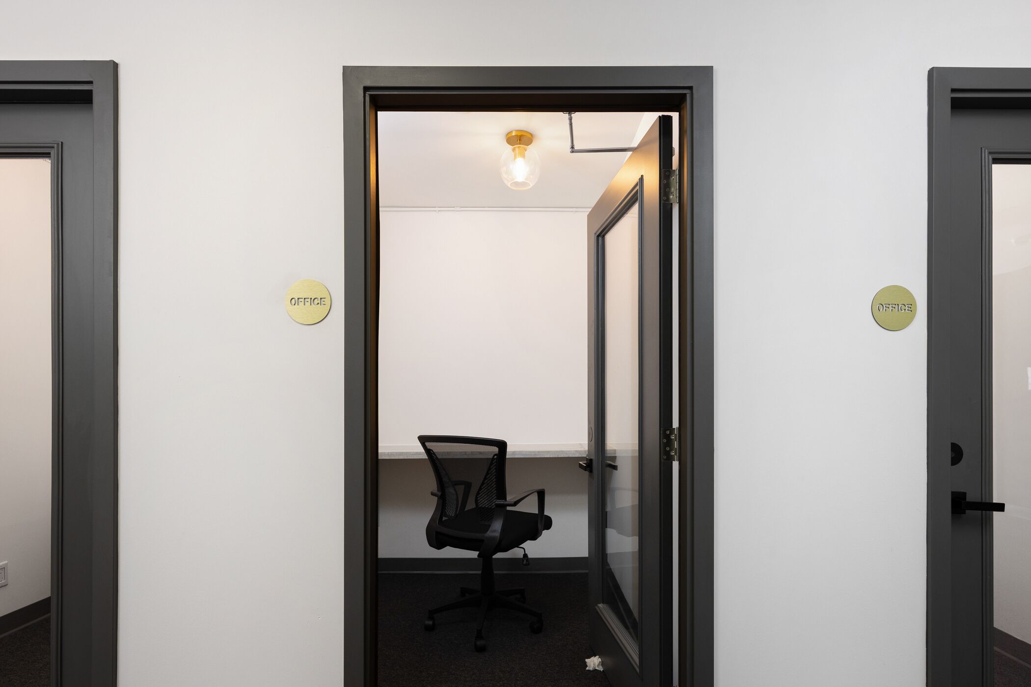 Ein schmaler Korridor mit zwei Türen, auf denen „OFFICE“ steht, der zu einem Raum mit einem einzelnen Bürostuhl und einer Deckenleuchte führt.