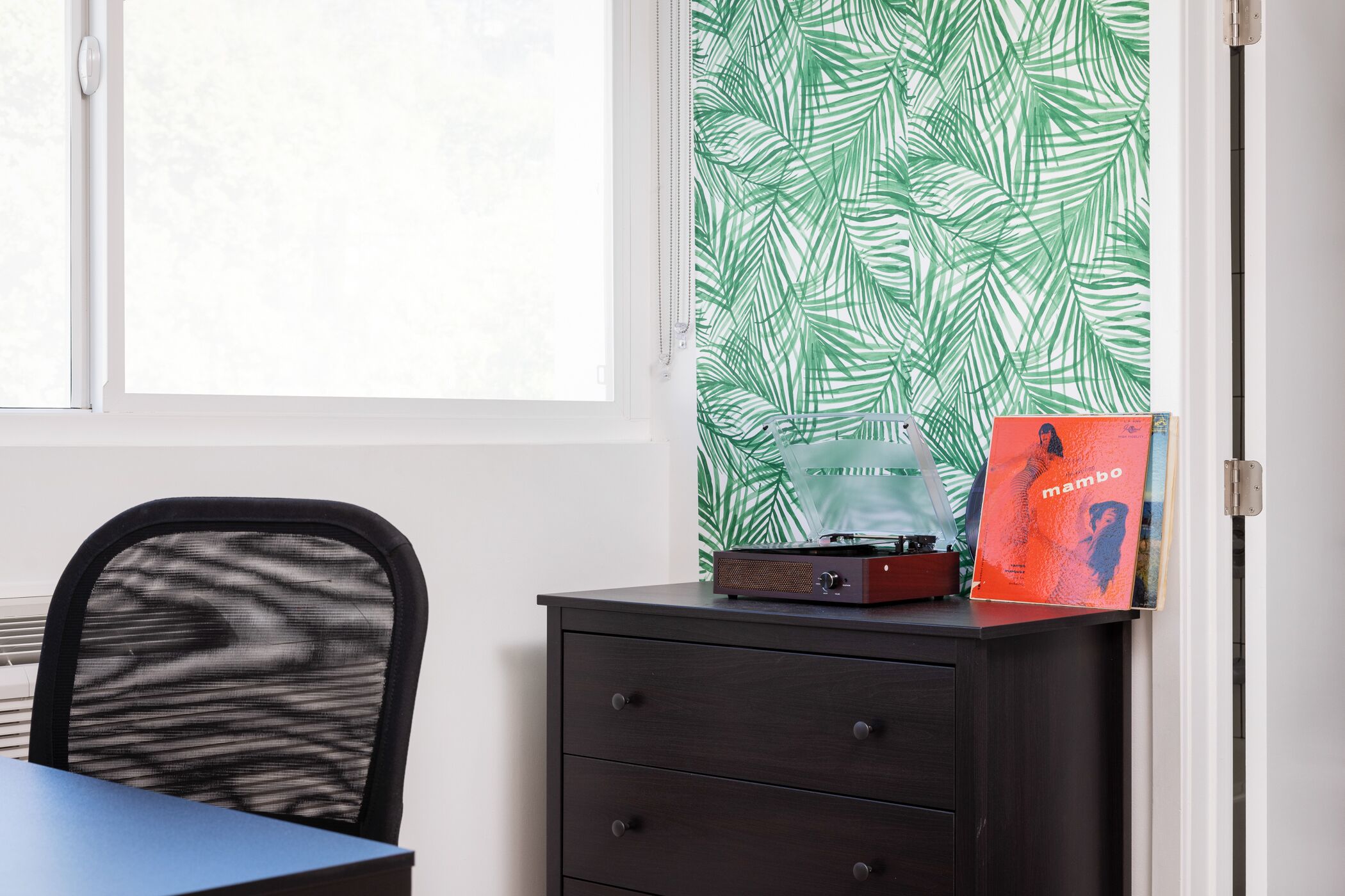 Una habitación con ventana, una silla, un escritorio, un aparador oscuro y una pared con estampado de hojas verdes.