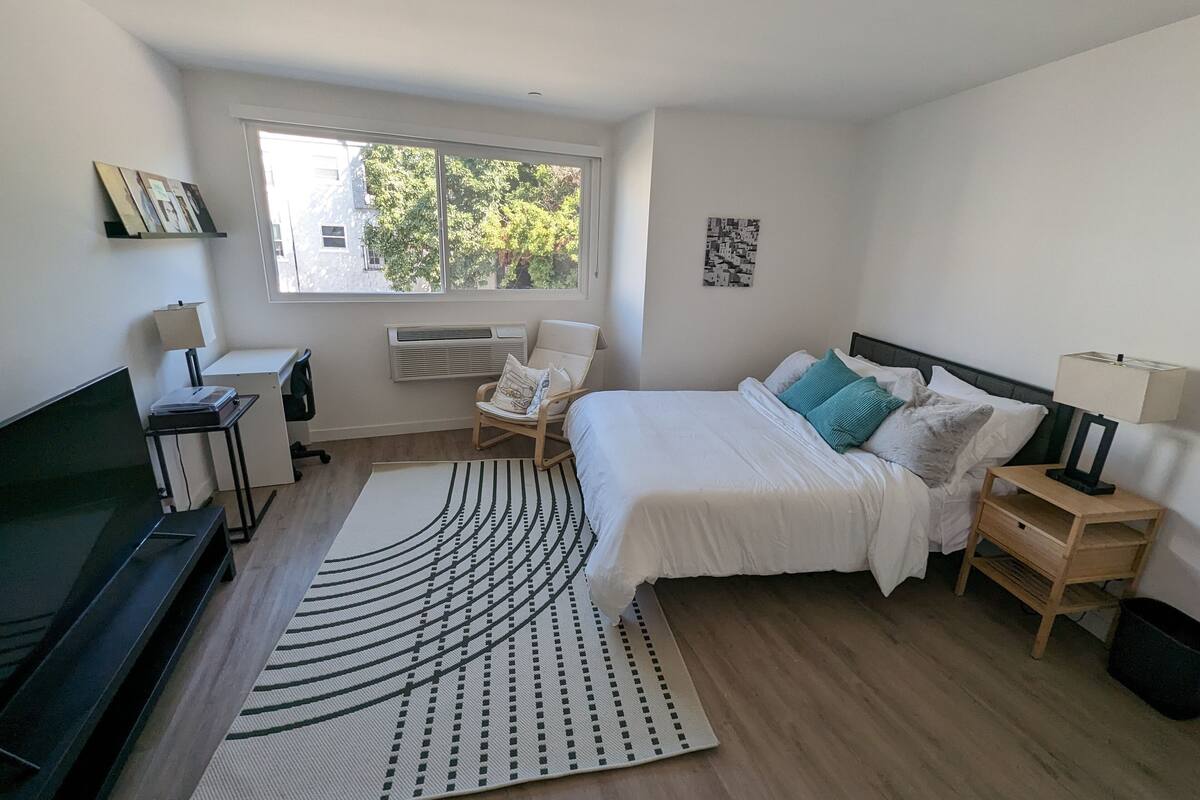 A modern bedroom with a large bed, a desk, a chair, a TV, and a window with a view of trees.