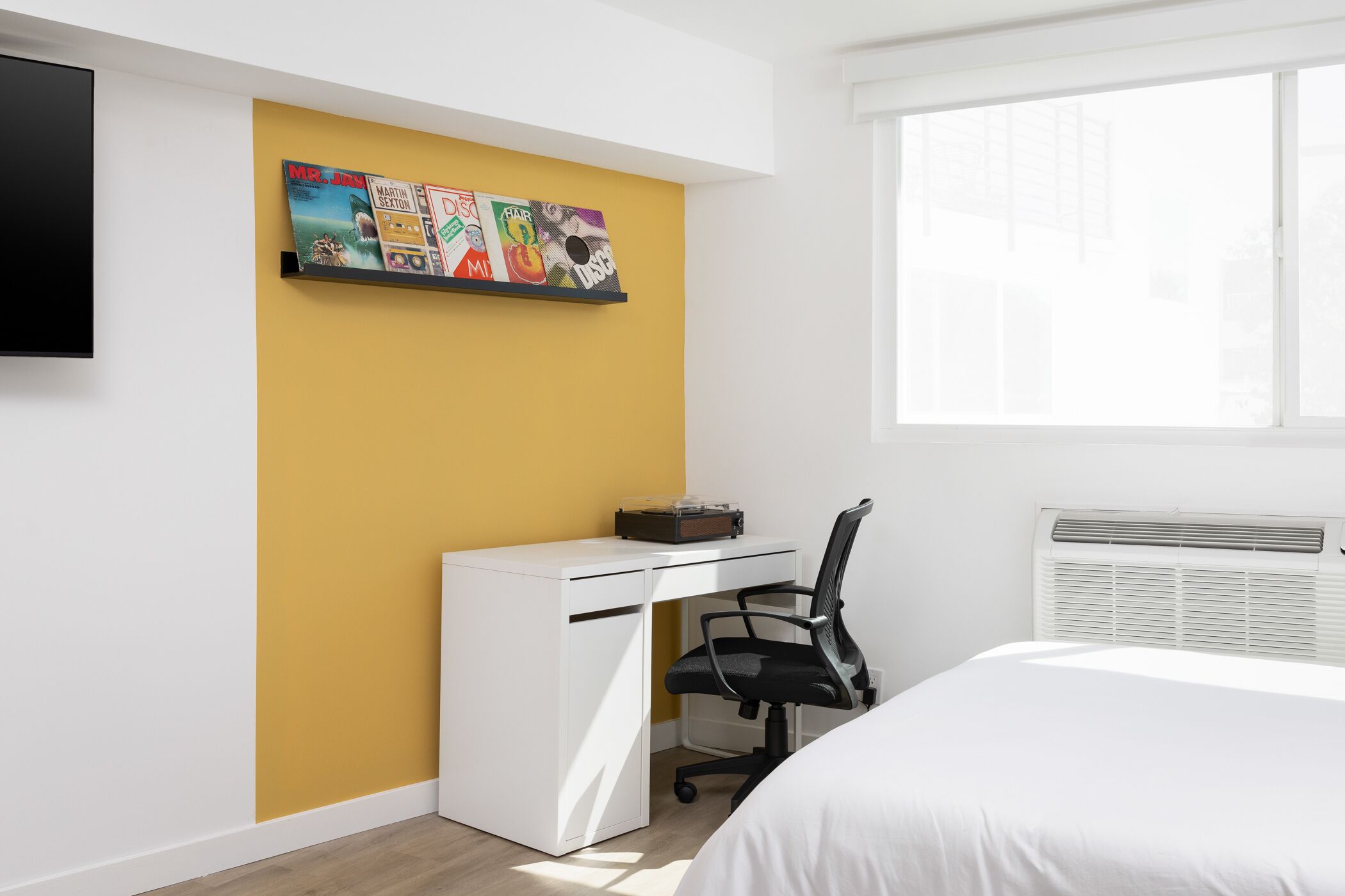 Un dormitorio moderno con una cama blanca, un escritorio con una silla, un televisor montado en la pared y un amplio ventanal.