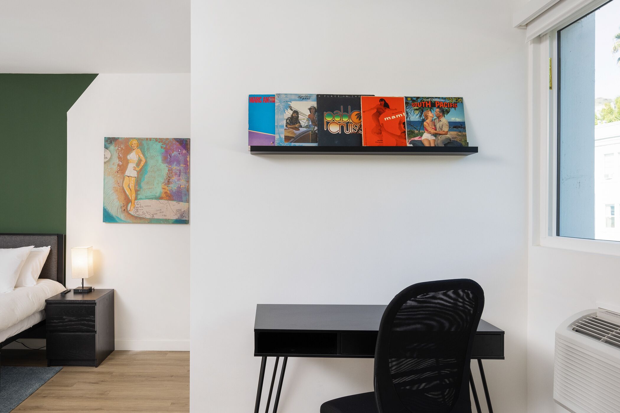 Une chambre moderne comprenant un lit, une table de chevet, une lampe, un bureau, une chaise et une œuvre d’art accrochée au mur.