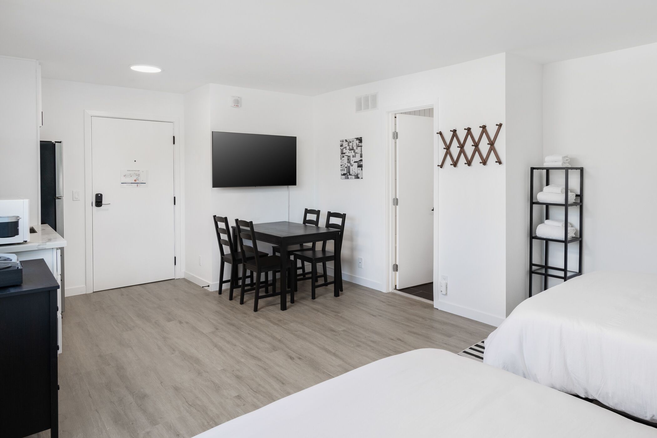 A modern hotel room with a bed, a desk with a computer, a dining table with chairs, and a TV mounted on the wall.