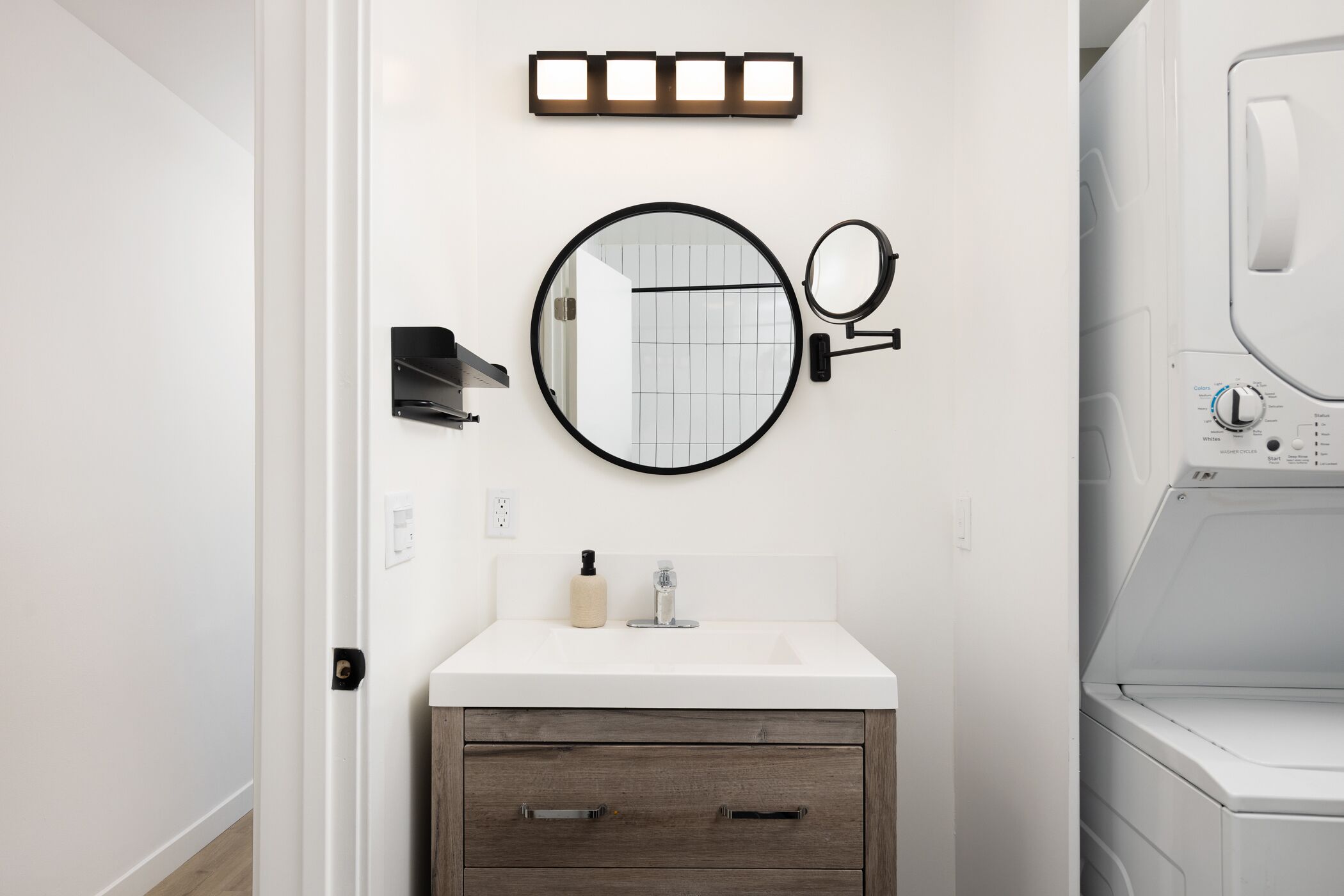 Un baño con un lavabo blanco, un espejo redondo, un dispensador de jabón y una lavadora.