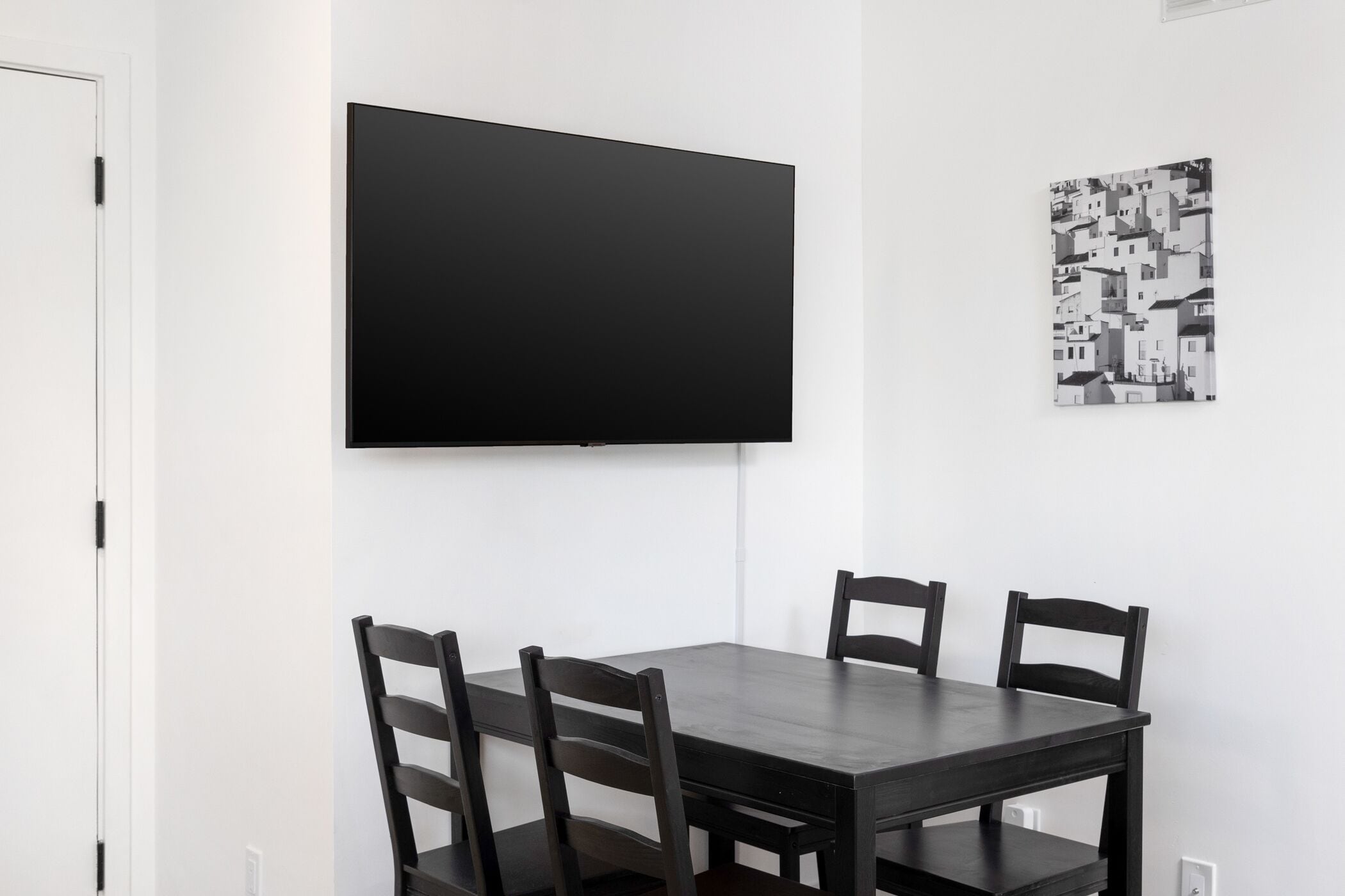 A dining area with a dark wooden table and four chairs, a flat-screen TV mounted on the wall, and a black and white patterned artwork on the wall.