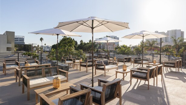 An outdoor patio with wooden furniture and large umbrellas, surrounded by buildings and palm trees. - Fenix Hotel Hollywood (Los Angeles)