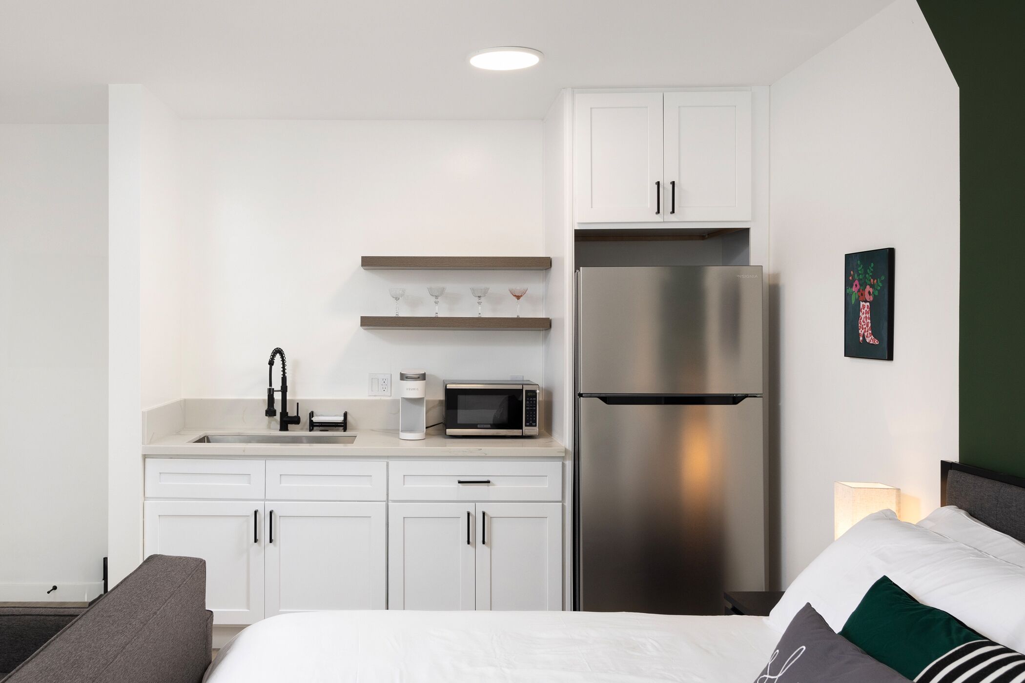 A modern hotel room with a kitchenette, a bed with pillows, and a nightstand with a lamp.