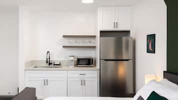 A modern hotel room with a kitchenette, a bed with pillows, and a nightstand with a lamp.