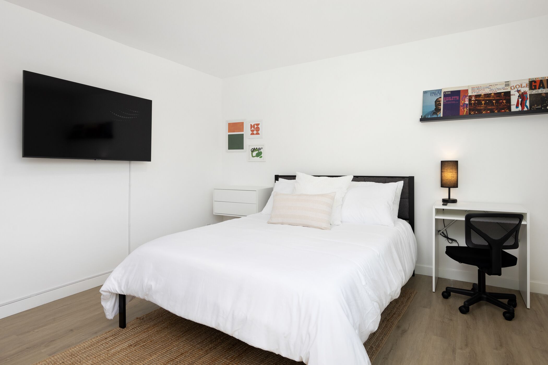 A modern bedroom with a bed, a desk, a chair, a TV, and wall art.