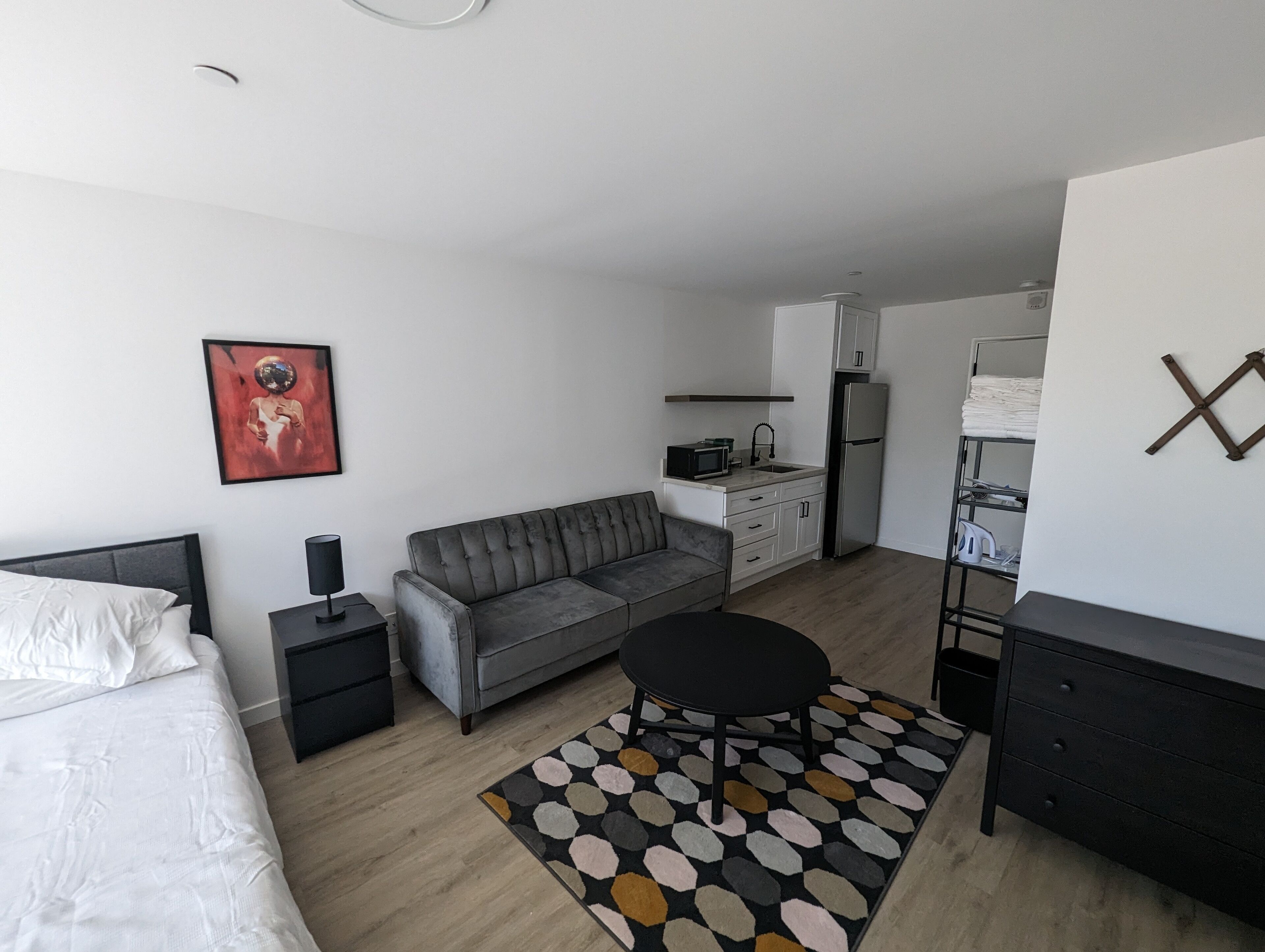 Un espacio habitable moderno y compacto con cama, sofá gris, pequeña mesa redonda y kitchenette.