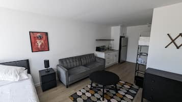 Un espacio habitable moderno y compacto con cama, sofá gris, pequeña mesa redonda y kitchenette.