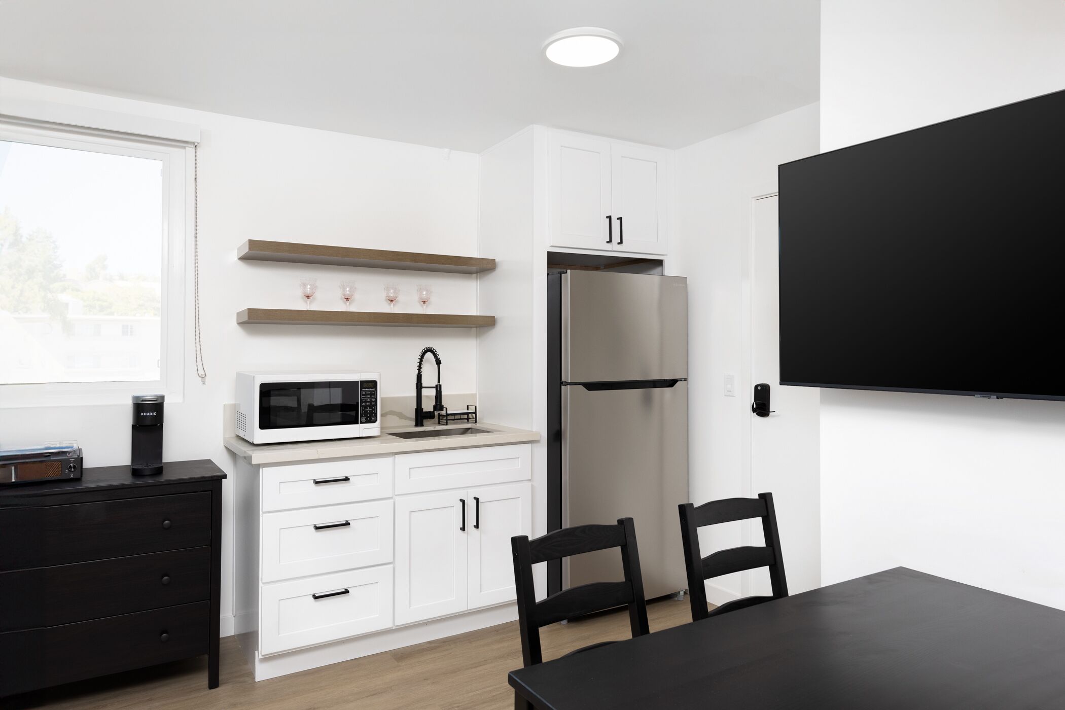 A modern kitchen with white cabinets, a stainless steel refrigerator, a microwave, and a black dining table with chairs.