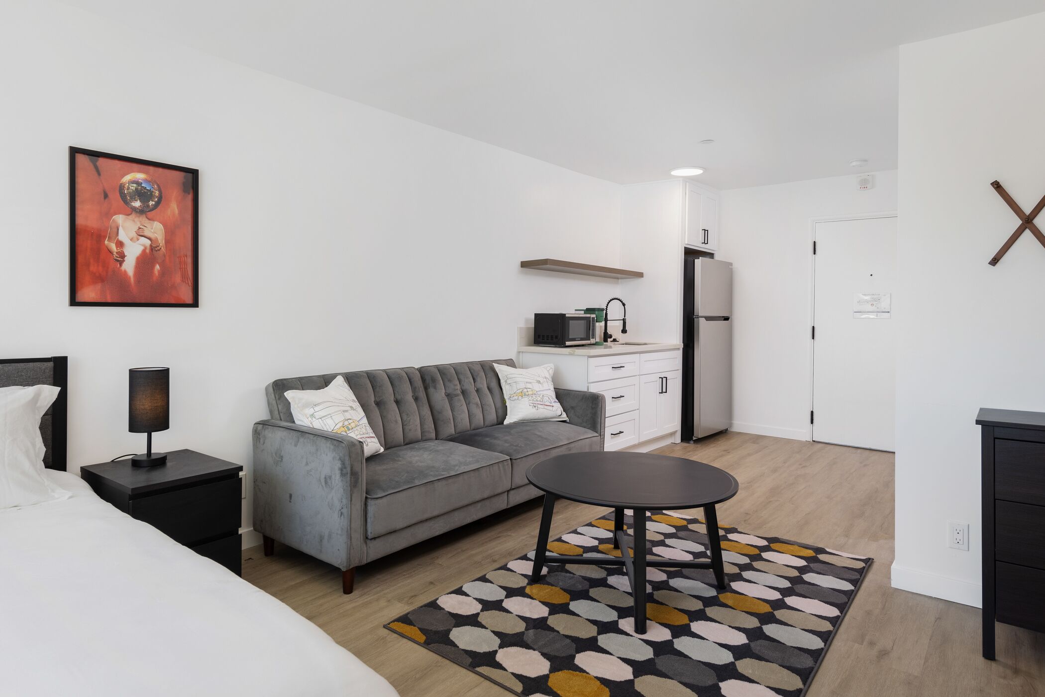 A modern hotel room with a grey sofa, a small round table, a bed, a nightstand, a lamp, a microwave, a refrigerator, and a painting on the wall.