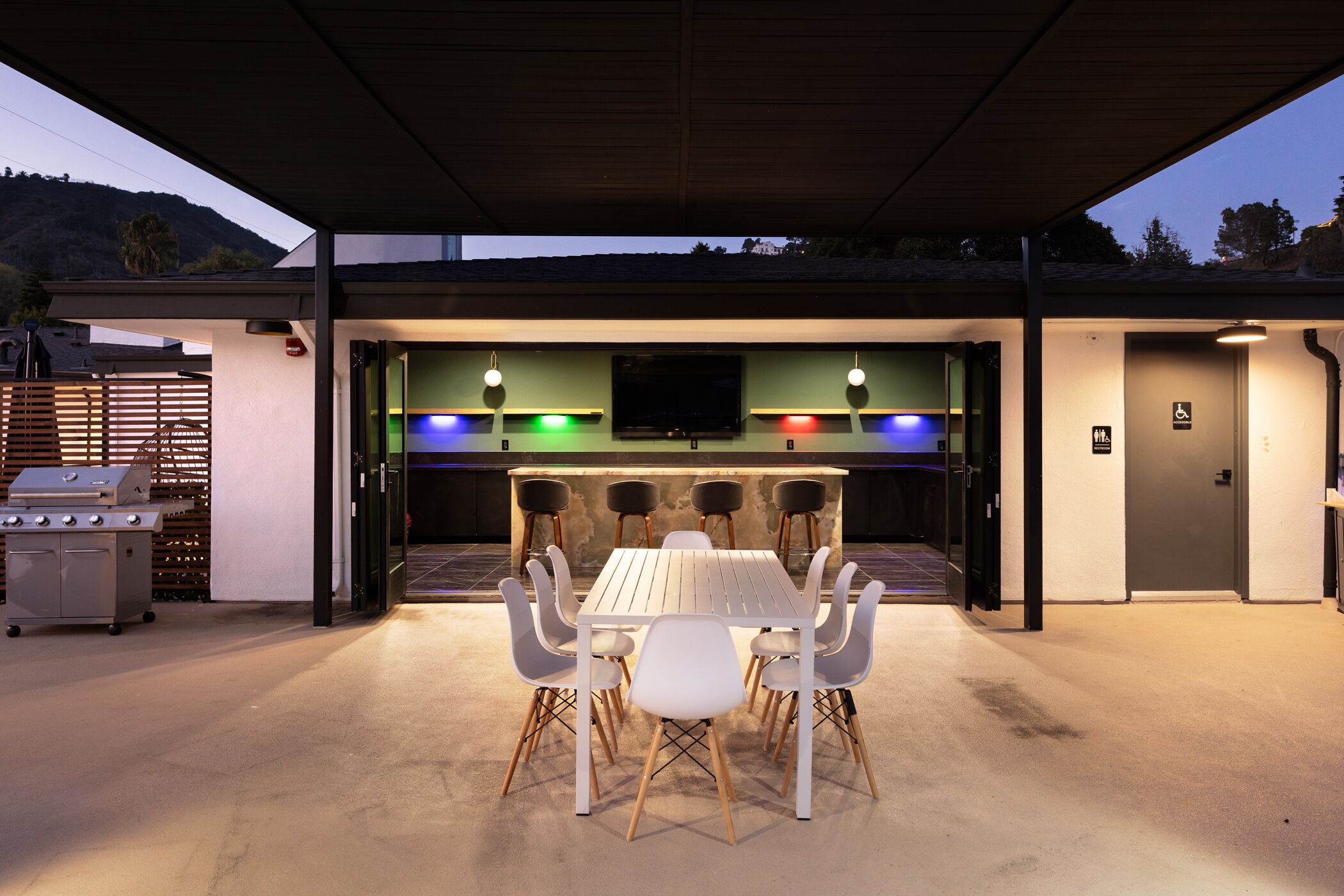 An outdoor dining area with a table and chairs, a grill, and a TV screen.