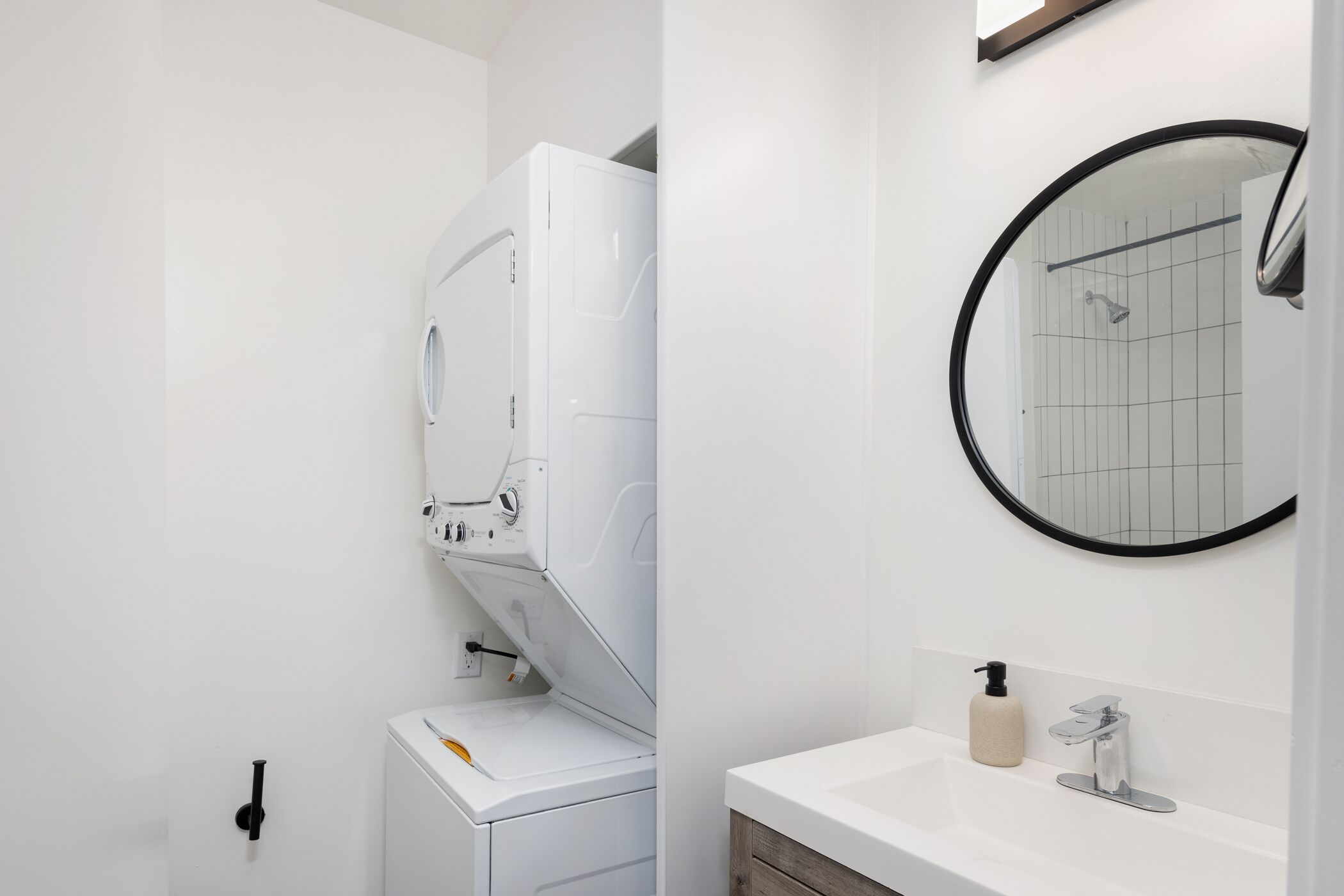Une salle de bain moderne équipée d’un lave-linge et d’un sèche-linge intégrés, d’un miroir rond, d’un lavabo avec un distributeur de savon et d’un porte-serviettes.