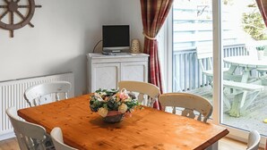 A wooden dining table with chairs, a television on a cabinet, and a view of the outside deck. - 3 bedroom accommodation in Pentraeth (Pentraeth)
