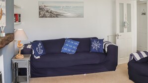 A living room with a navy blue sofa, a painting of a coastal scene, a white door, and a small table with a lamp. - 3 bedroom accommodation in Pentraeth (Pentraeth)