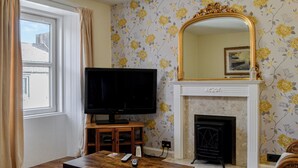 A living room with a television on a wooden stand, a fireplace, and floral wallpaper. - 3 bedroom accommodation in Castle Douglas (Castle Douglas)