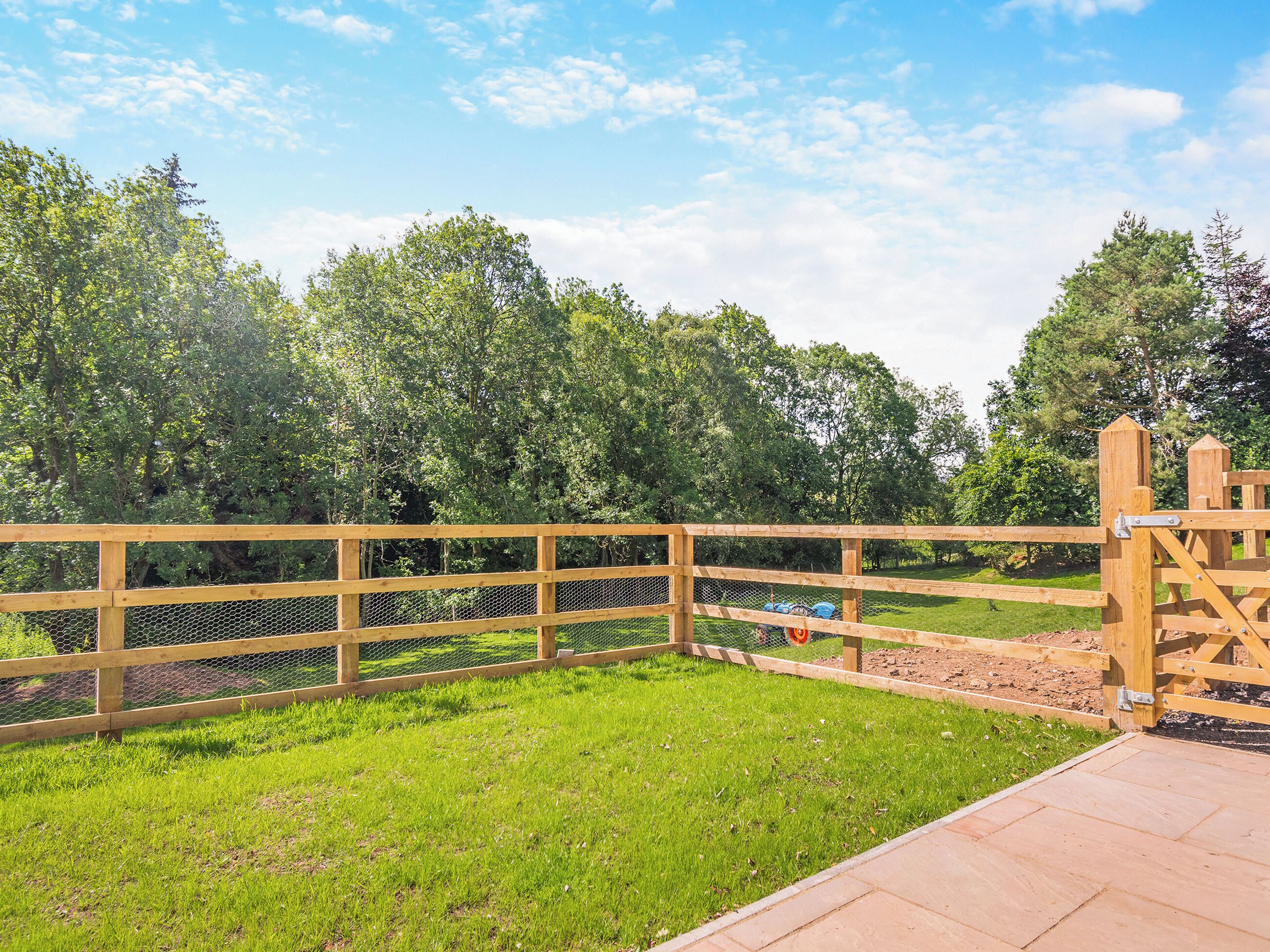 A fenced outdoor area with a wooden gate, a grassy field, and trees in the background.