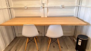 Une pièce de taille modeste, avec un bureau en bois et deux chaises blanches, une poubelle noire et une porte blanche.