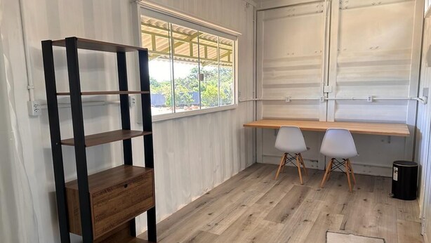 Une pièce avec un plancher en bois, une étagère en métal, deux chaises blanches, un bureau et une fenêtre donnant sur un paysage verdoyant.