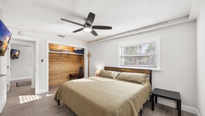Un dormitorio con cama, ventilador de techo, ventana con persianas, pared de madera y puerta que da a otra habitación.