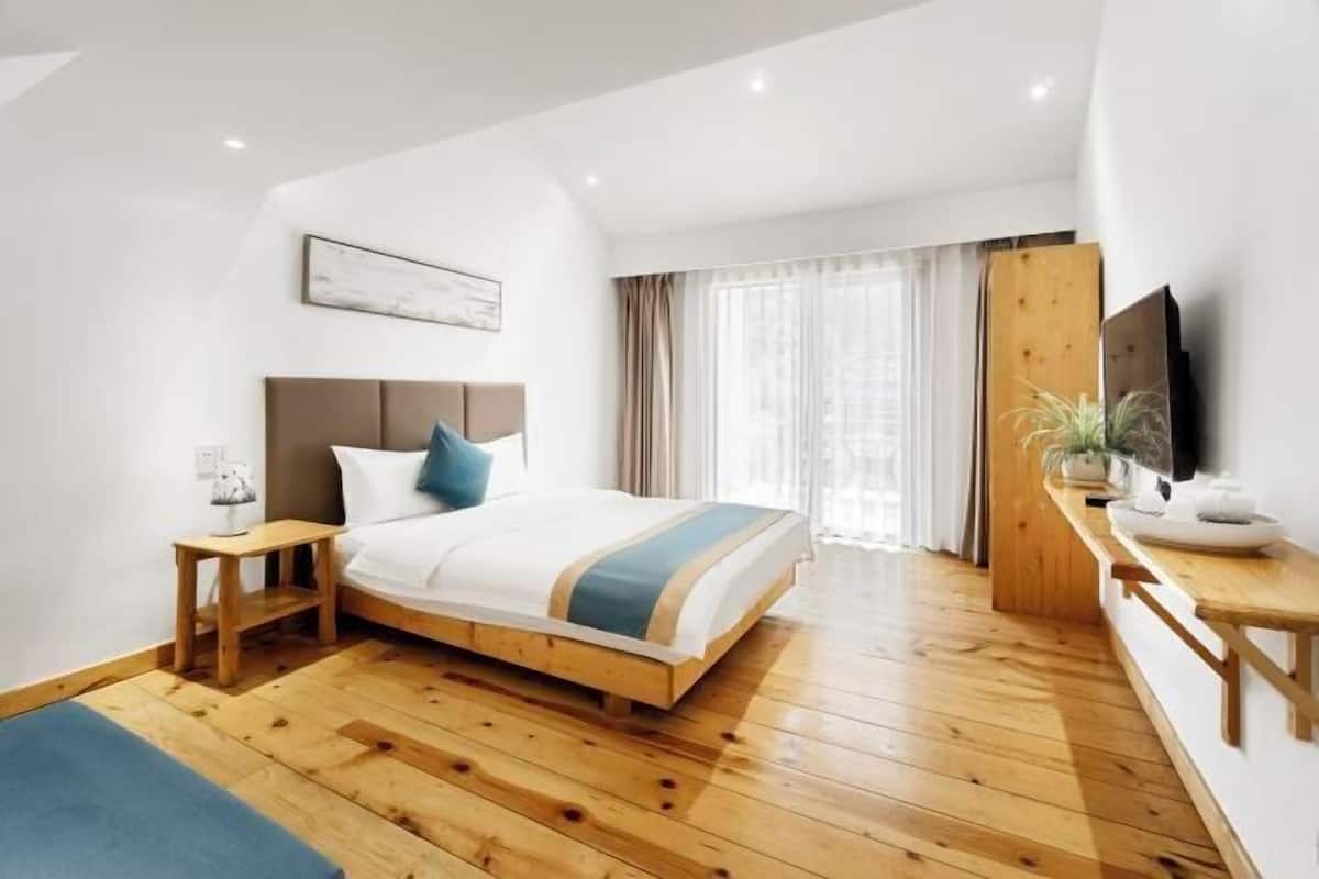 Un dormitori amb sòl de fusta, un llit amb edredó blau i blanc a ratlles, una nitzeta, un escriptori de fusta, una televisió i una finestra amb cortines.