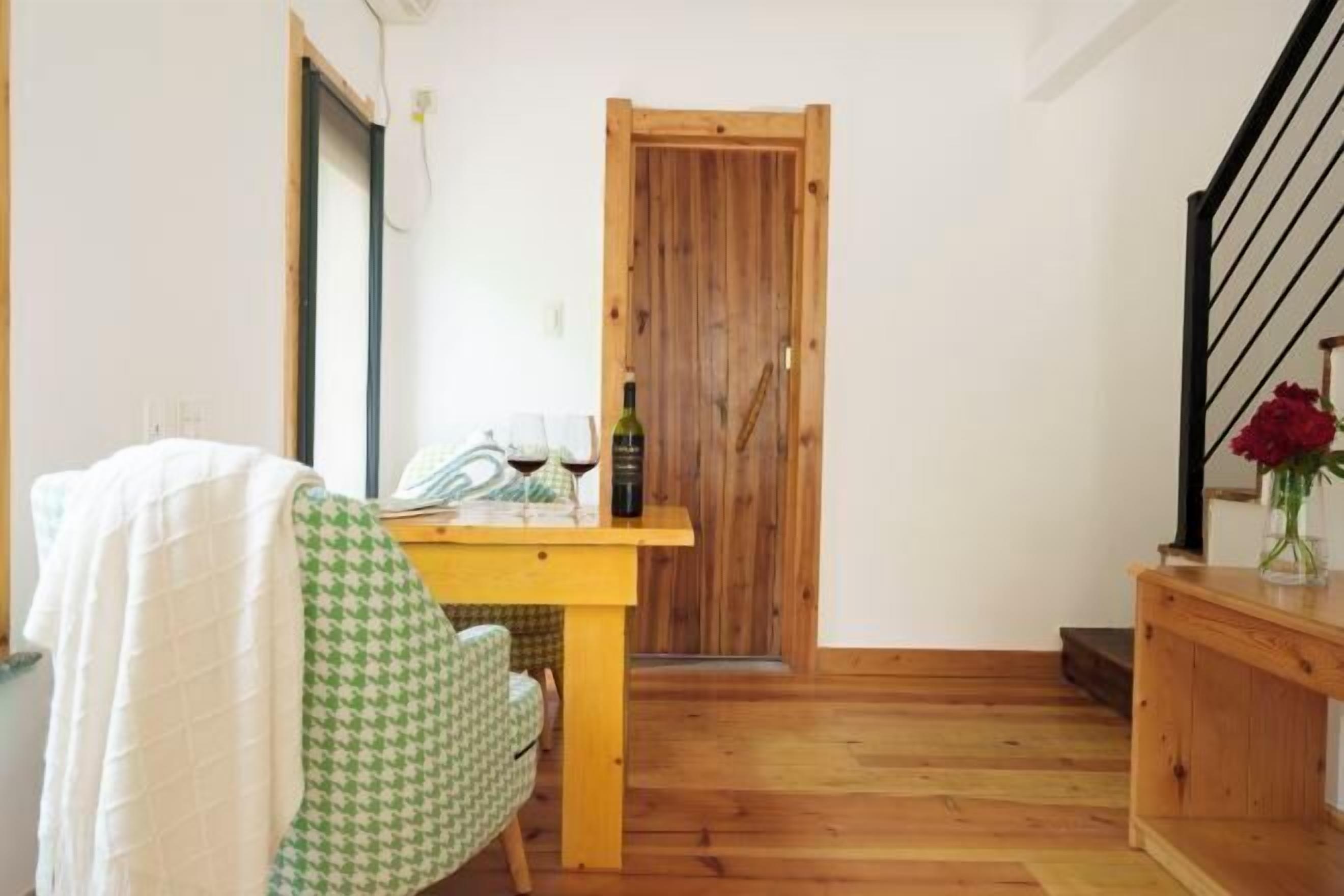 A small, cozy room with a wooden door, a dining table with a bottle and glasses, a chair, and a staircase with a vase of flowers.
