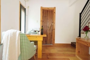 A small, cozy room with a wooden door, a dining table with a bottle and glasses, a chair, and a staircase with a vase of flowers.