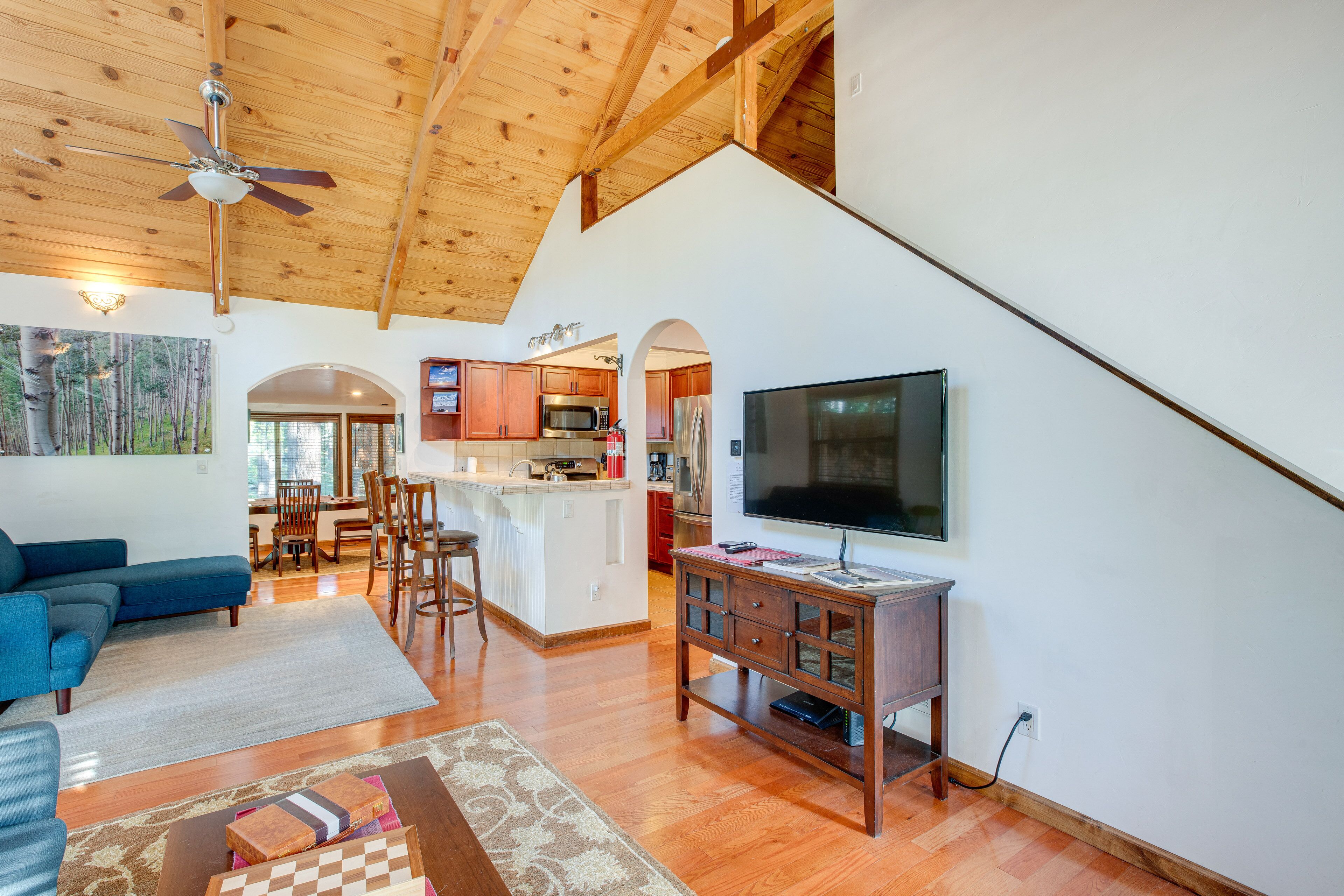 Un'ampia zona giorno con divano blu, pavimento in legno, ventilatore a soffitto, televisore a schermo piatto e cucina dotata di elettrodomestici moderni.