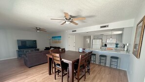 Un grand espace de vie comprenant une table à manger en bois, deux ventilateurs de plafond, un téléviseur à écran plat, un canapé et une cuisine équipée d’un réfrigérateur et d’un four à micro-ondes.