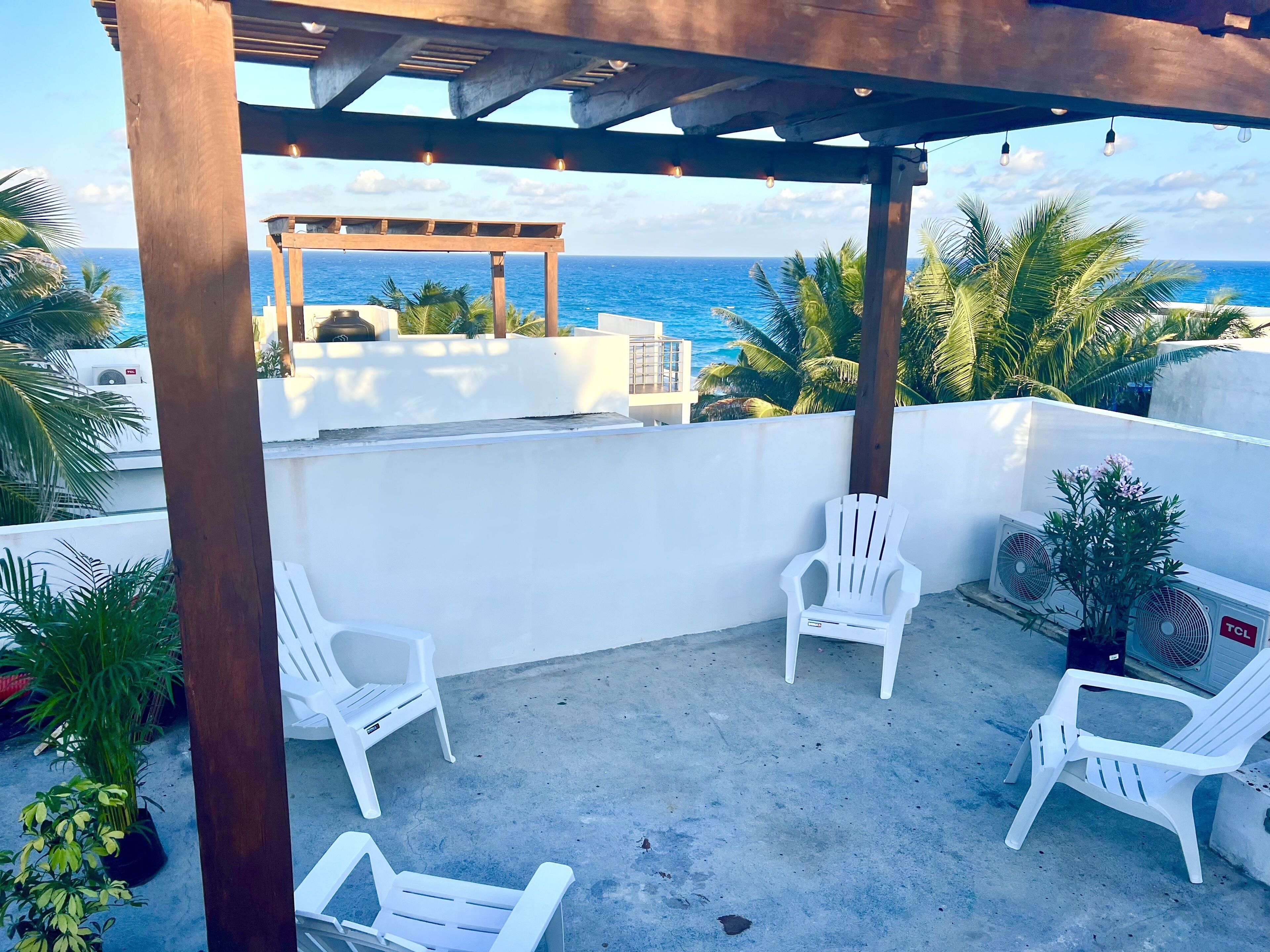 A covered outdoor seating area with white plastic chairs, a pergola, and a view of the ocean.