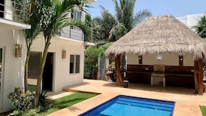 Una casa moderna con piscina, una estructura de techo de paja y abundante follaje.
