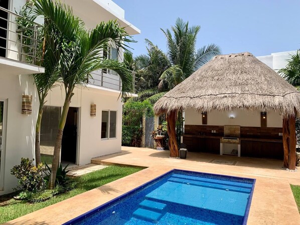 Una casa moderna con piscina, una estructura de techo de paja y abundante follaje.