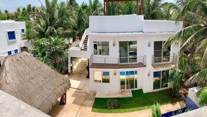 Una casa moderna de dos pisos con piscina, rodeada de palmeras y una estructura con techo de paja.